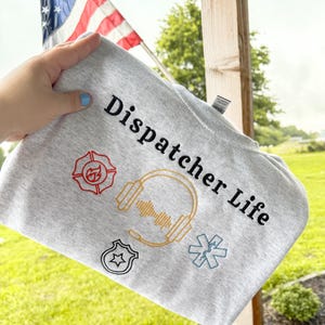 May include: A light gray sweatshirt with the words "Dispatcher Life" in black script. The sweatshirt features embroidered symbols of a fire department emblem, headphones, a police badge, and an emergency medical services star.