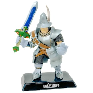May include: A Final Fantasy IX figurine of a knight in silver armor with a brown strap, wearing a helmet with a feather. The knight holds a sword with a blue blade and a green hilt. The figure stands on a black base labeled Final Fantasy IX.