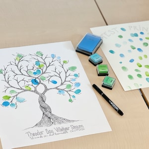 K&ouml;nnte beinhalten: Ein wei&szlig;es Blatt Papier mit einer schwarzen und wei&szlig;en Baumzeichnung. Der Baum hat gr&uuml;ne und blaue Fingerabdr&uuml;cke. Der Text "Theodor Bay Villekier Skoum Til minde om din barned&aring;b 02.07.2022" befindet sich am unteren Rand der Seite. Auf dem Tisch liegen vier Stempelkissen und ein schwarzer Stift.