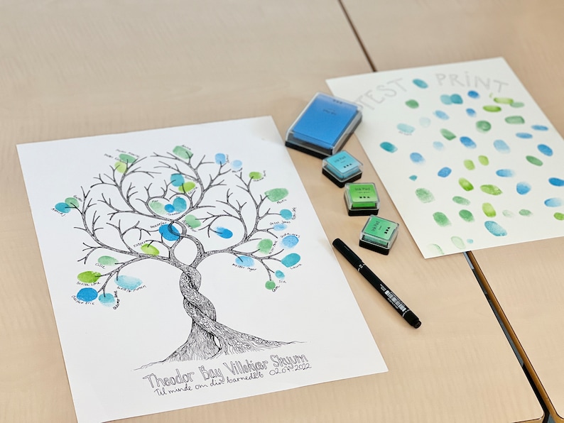 K&ouml;nnte beinhalten: Ein wei&szlig;es Blatt Papier mit einer schwarzen und wei&szlig;en Baumzeichnung. Der Baum hat gr&uuml;ne und blaue Fingerabdr&uuml;cke. Der Text "Theodor Bay Villekier Skoum Til minde om din barned&aring;b 02.07.2022" befindet sich am unteren Rand der Seite. Auf dem Tisch liegen vier Stempelkissen und ein schwarzer Stift.
