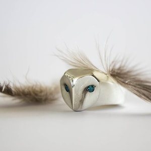 May include: A silver ring with a detailed owl head design. The owl's eyes are set with turquoise gemstones. A single brown feather is attached to the ring.