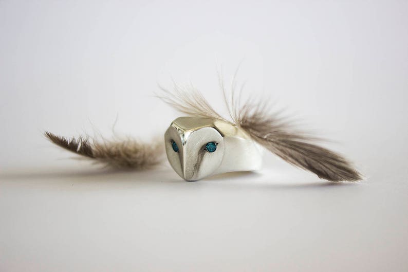 May include: A silver ring with a detailed owl head design. The owl's eyes are set with turquoise gemstones. A single brown feather is attached to the ring.