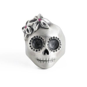 Puede incluir: Un anillo de calavera de plata con un diseño detallado de calavera de azúcar. La calavera tiene dos ojos negros con puntos blancos alrededor y una boca cosida. La calavera está coronada con tres flores plateadas con piedras preciosas rojas en el centro.