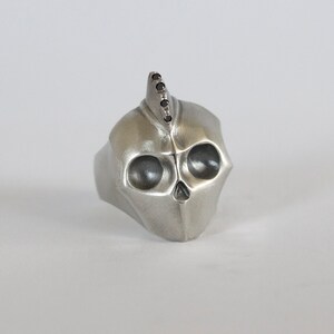 Könnte beinhalten: Ein silberner Totenkopfring mit einem schwarzen Diamanten-Mohawk. Der Totenkopf hat ein detailliertes Design mit hohlen Augenhöhlen.