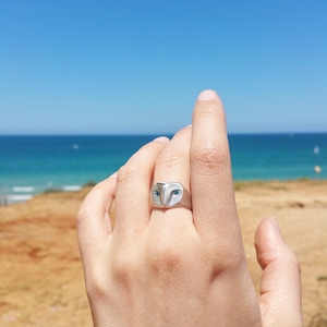 May include: A silver ring with an owl design. The owl's eyes are blue gemstones. The ring is worn on a person's finger against a background of a sandy beach and blue ocean.