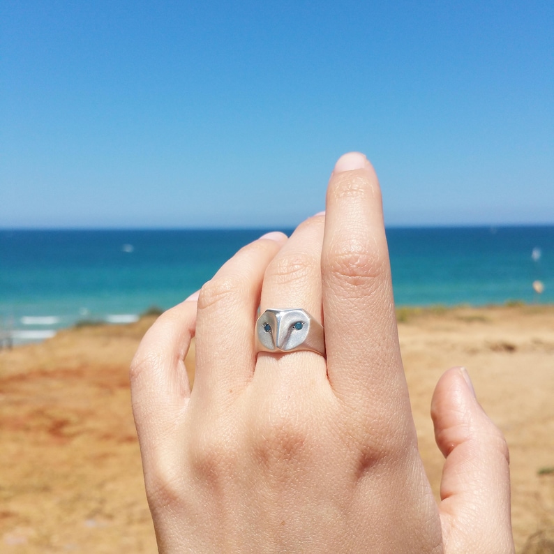 May include: A silver ring with an owl design. The owl's eyes are blue gemstones. The ring is worn on a person's finger against a background of a sandy beach and blue ocean.