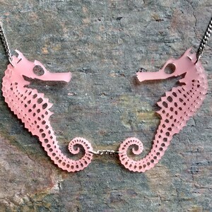 May include: A pink seahorse necklace with two seahorses facing each other. The seahorses are connected by a silver chain.