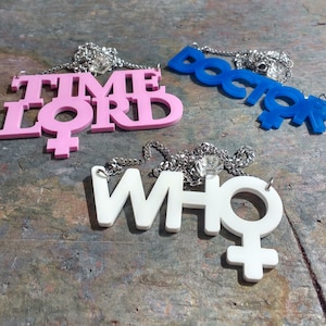 May include: Three necklaces with pendants featuring the words "TIME LORD", "DOCTOR", and "WHO" in pink, blue, and white, respectively. Each pendant has a stylized female symbol.