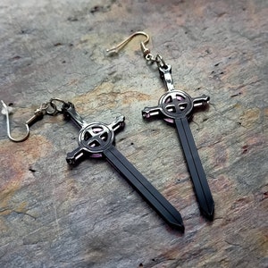 Könnte beinhalten: Ein Paar schwarze Schwertohrringe mit einem rosafarbenen Kreuzdesign. Die Ohrringe sind aus Metall und haben einen Hakenverschluss.
