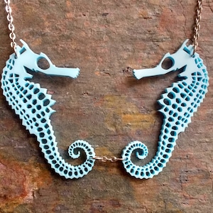 May include: A light blue necklace featuring two seahorse pendants with a delicate chain. The seahorses are intricately designed with a patterned texture.