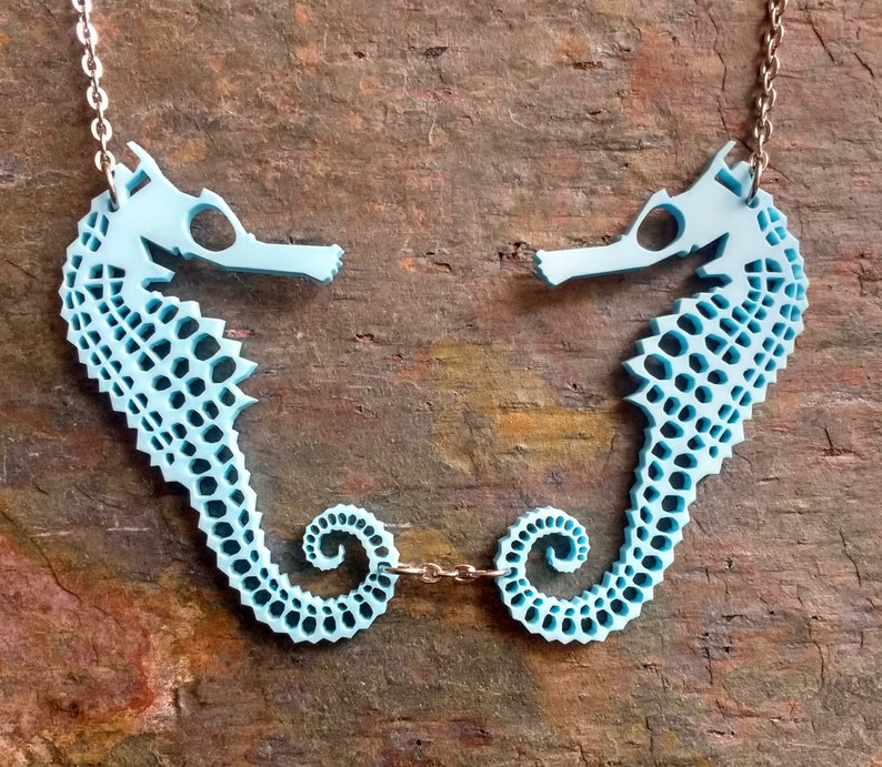 May include: A light blue necklace featuring two seahorse pendants with a delicate chain. The seahorses are intricately designed with a patterned texture.