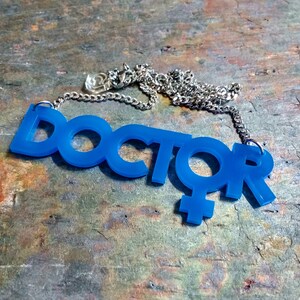May include: A blue acrylic necklace with the word "DOCTOR" and a female symbol. The necklace is on a silver chain.