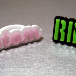 May include: Two decorative accessories. One is a white ring with the word "Custom" in pink lettering. The other is a black rectangle with the word "RINGS" in green lettering.