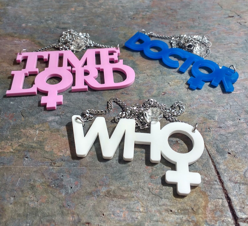 May include: Three necklaces with pendants featuring the words "TIME LORD", "DOCTOR", and "WHO" in pink, blue, and white, respectively. Each pendant has a stylized female symbol.