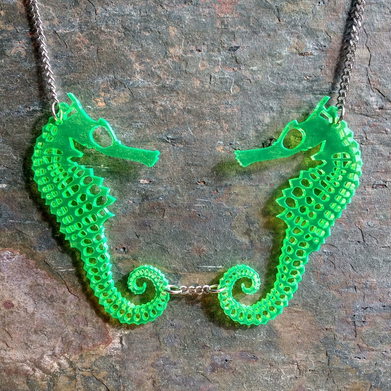 May include: A green necklace with two seahorse pendants connected by a silver chain. The seahorses are facing each other and have a detailed, textured design.