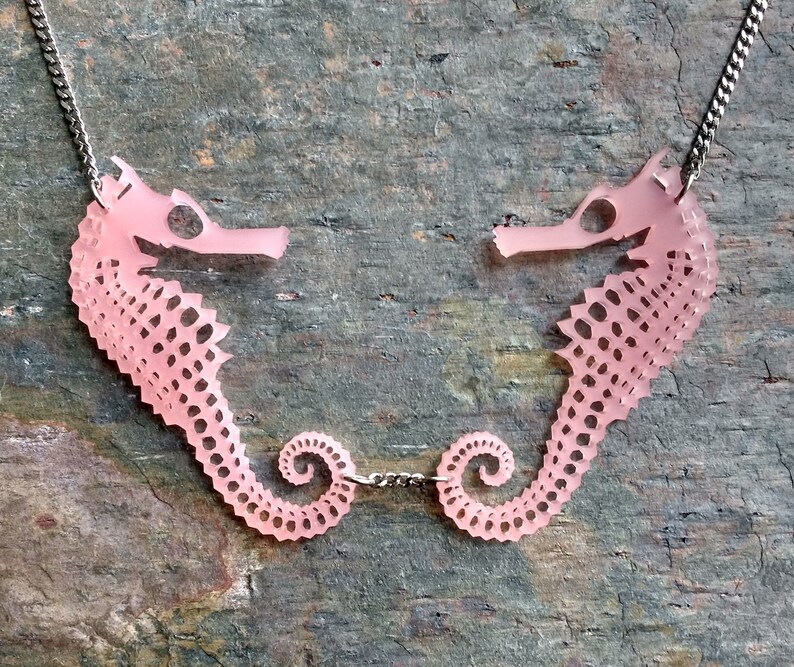 May include: A pink seahorse necklace with two seahorses facing each other. The seahorses are connected by a silver chain.