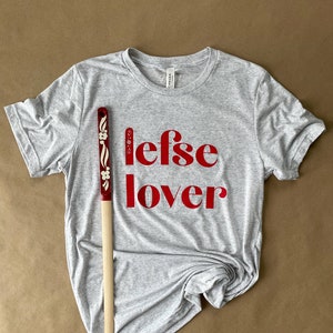 Peut inclure: Un t-shirt gris avec le texte "lefse lover" imprimé en rouge. Un rouleau à pâtisserie en bois avec des motifs floraux rouges et blancs est posé sur le t-shirt.