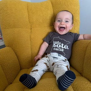 May include: A baby boy wearing a gray onesie with the text "little sota cutie" and a white outline of the state of Minnesota. He is sitting in a yellow armchair and smiling.