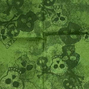 May include: Green fabric with a repeating pattern of dark green skulls and crossbones. The fabric has a mottled, distressed appearance. The skulls are detailed with eye sockets, noses, and teeth. The fabric is likely used for crafting or sewing projects.