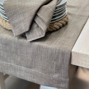 May include: A brown linen table runner with a rope trim. The runner is folded and placed on a table with a stack of plates on top.