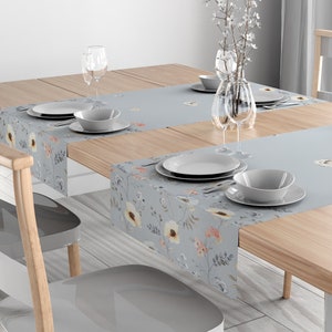May include: A light blue floral table runner with a delicate floral pattern. The runner is placed on a wooden table with two place settings, including plates, bowls, and glasses.