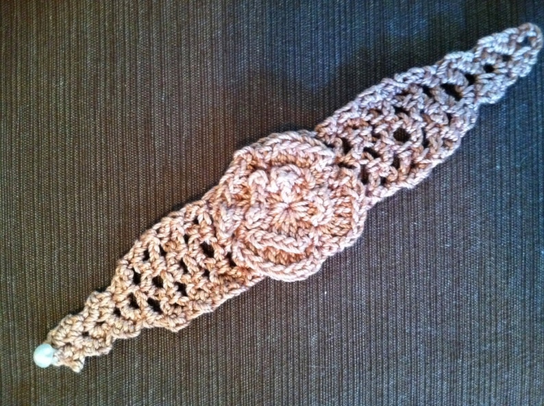 Puede incluir: Un brazalete de ganchillo con un dise&ntilde;o de flor en color marr&oacute;n claro. El brazalete tiene una cuenta blanca en el extremo.