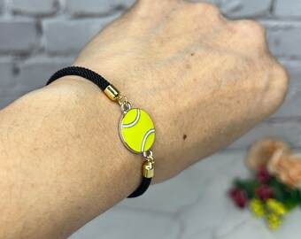 May include: A black cord bracelet with a gold charm featuring a yellow tennis ball.