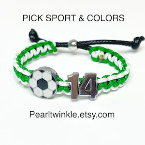 May include: Green and white braided bracelet with a silver soccer ball charm and a silver charm with the number 14. The bracelet has a black cord with a silver bead.