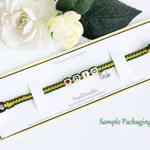 May include: A green and yellow friendship bracelet with the name "RIO" spelled out in white beads. A red heart bead is before the "R" and a round charm with the year "2016" is attached to the end of the bracelet. The bracelet is displayed on a white card with the text "Get Inspired. Go Inspire!" and "Pearl Twinkle Handcrafted Custom Jewelry". The card also says "Sample Packaging".