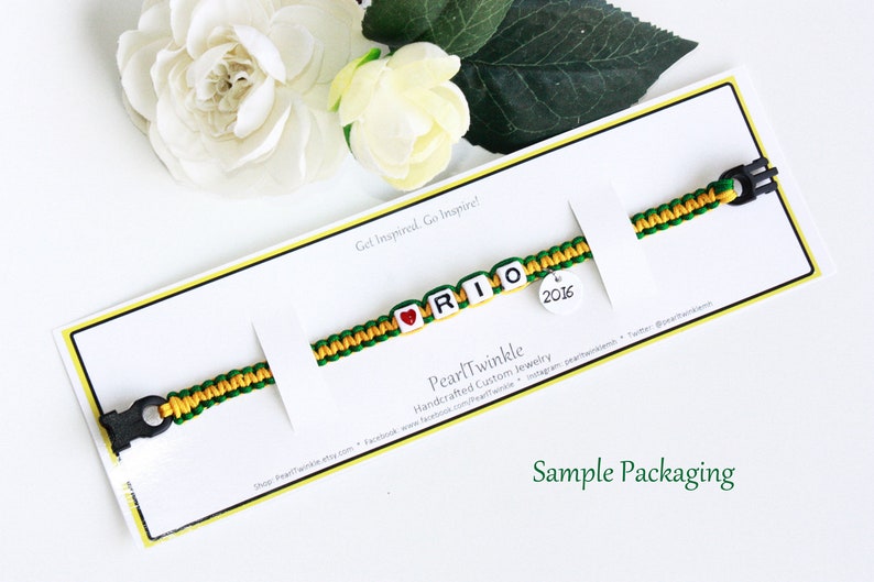 May include: A green and yellow friendship bracelet with the name "RIO" spelled out in white beads. A red heart bead is before the "R" and a round charm with the year "2016" is attached to the end of the bracelet. The bracelet is displayed on a white card with the text "Get Inspired. Go Inspire!" and "Pearl Twinkle Handcrafted Custom Jewelry". The card also says "Sample Packaging".