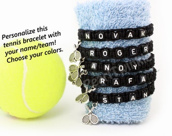 May include: Five black tennis bracelet charms with white letters spelling out the names NOVAK, ROGER, ANDY, RAFA, and STAN. Each charm has a silver tennis racket pendant.