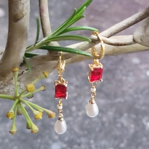 Può includere: Orecchini a cerchio dorati con una pietra quadrata rossa e una perla bianca che pendono dal fondo.