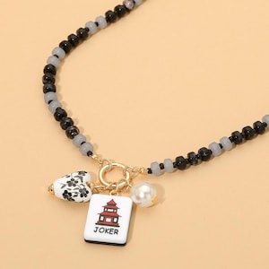 May include: A beaded necklace featuring alternating black and translucent gray beads. A gold-toned circular clasp holds three charms: a white heart with black floral patterns, a white rectangular tile with a red pagoda and the text JOKER, and a pearl.