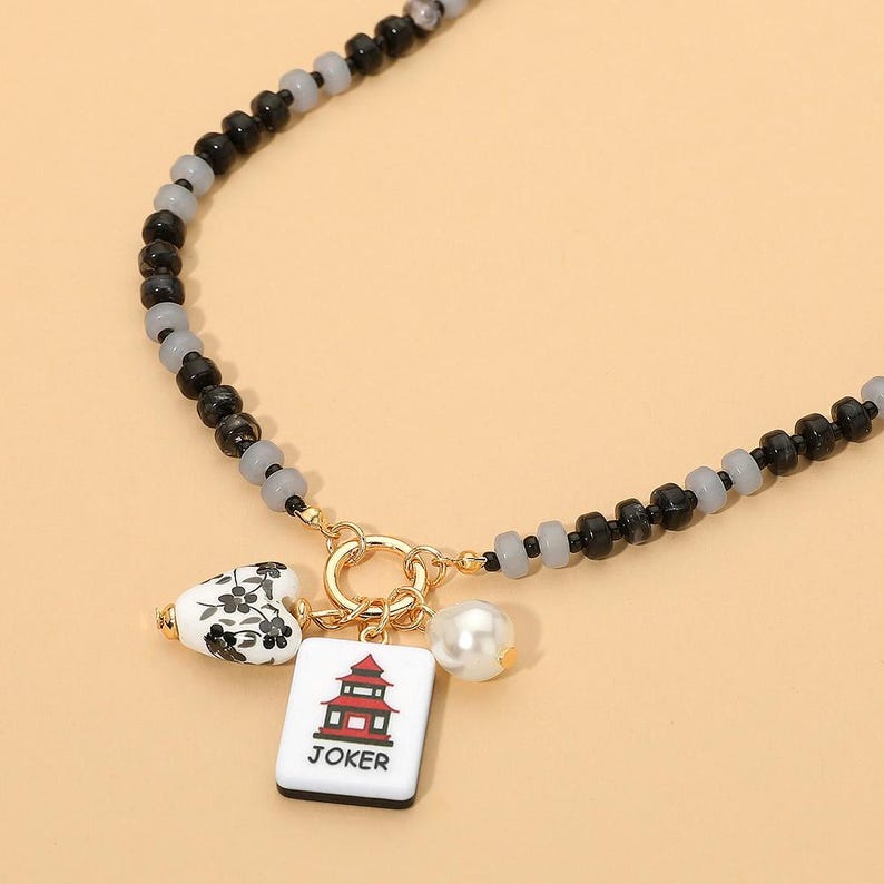 May include: A beaded necklace featuring alternating black and translucent gray beads. A gold-toned circular clasp holds three charms: a white heart with black floral patterns, a white rectangular tile with a red pagoda and the text JOKER, and a pearl.