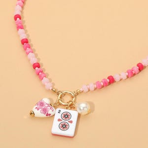 May include: A beaded necklace featuring a strand of pink and red beads with a gold-toned circular clasp. Three charms hang from the clasp: a white heart with pink floral patterns, a white rectangular mahjong tile with the number 2, and a white pearl.
