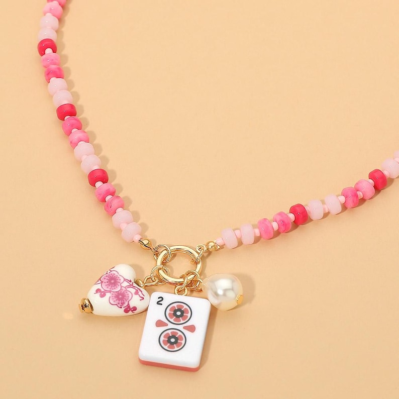 May include: A beaded necklace featuring a strand of pink and red beads with a gold-toned circular clasp. Three charms hang from the clasp: a white heart with pink floral patterns, a white rectangular mahjong tile with the number 2, and a white pearl.