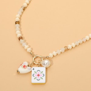 May include: A beaded necklace featuring a series of off-white and translucent brown beads. The necklace includes a gold-toned metal ring clasp holding three charms: a white heart with a floral pattern, a white pearl, and a white mahjong tile with a number 4.