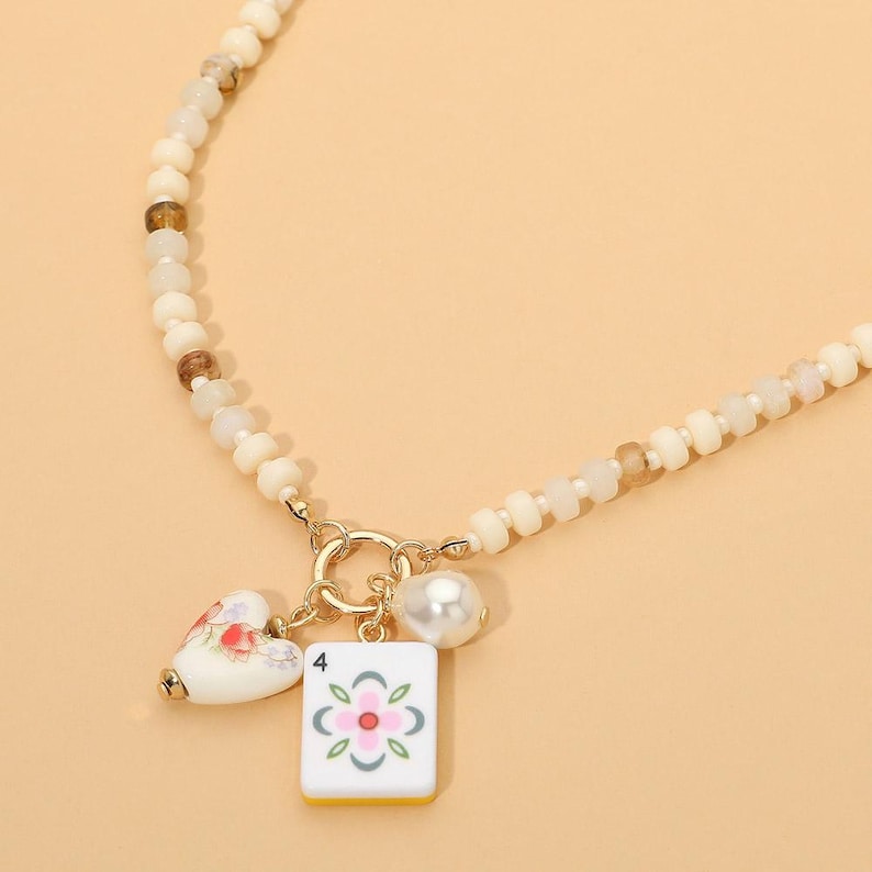 May include: A beaded necklace featuring a series of off-white and translucent brown beads. The necklace includes a gold-toned metal ring clasp holding three charms: a white heart with a floral pattern, a white pearl, and a white mahjong tile with a number 4.