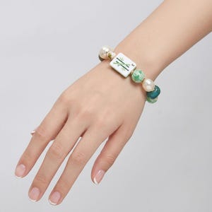 May include: A stretch bracelet featuring a rectangular white mahjong tile with a green bamboo design, interspersed with round white faux pearls, marbled green beads, and solid dark teal beads on an elastic cord.