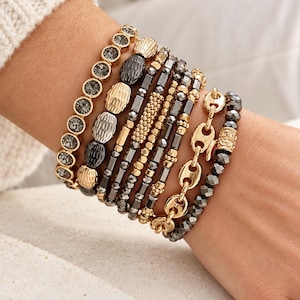 May include: A stack of seven bracelets featuring a mix of dark gray faceted beads, gold-toned metallic beads, and a gold-toned chain link bracelet. The set includes various textures such as smooth, ribbed, and faceted surfaces in a layered design.