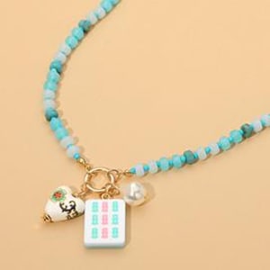 May include: A beaded necklace featuring a strand of light blue and turquoise beads. The necklace includes a gold-toned circular clasp with three charms: a white heart with a floral design, a white rectangular mahjong tile, and a white pearl bead.