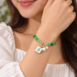 May include: A beaded bracelet featuring various shades of green beads, a gold-toned circular clasp, and three charms: a white rectangular tile with a green bamboo graphic, a small white round bead, and a larger white pearl-like bead.