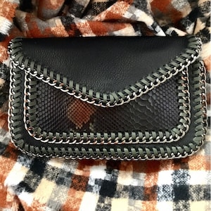 May include: Black clutch with a snakeskin pattern on the front flap. The clutch features a silver chain trim and a matching black leather trim. The bag is rectangular and has a fold-over flap closure.