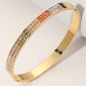 May include: Gold-tone stainless steel bangle bracelet with two rows of colorful square-cut gemstones. The bracelet features a hinged clasp and the words "Stainless Steel" are engraved on the side.