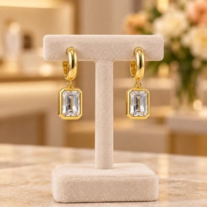 May include: A pair of gold hoop earrings featuring rectangular emerald-cut clear stones suspended from each hoop. The earrings are displayed on a beige T-shaped stand, showcasing the polished gold metal finish and the faceted clarity of the stones.