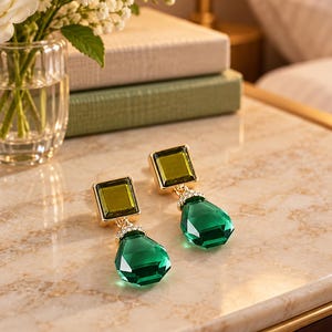 May include: A pair of drop earrings featuring a square olive green stone set in gold-tone metal, connected to a faceted emerald green teardrop-shaped stone. The connection point is accented with a small row of clear white crystals.