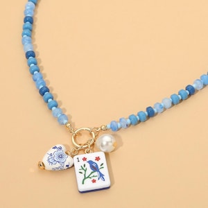 May include: A beaded necklace featuring various shades of blue beads and a gold-toned circular clasp. Three charms hang from the clasp: a white heart with blue floral patterns, a white rectangular mahjong tile with a blue bird, and a white pearl.