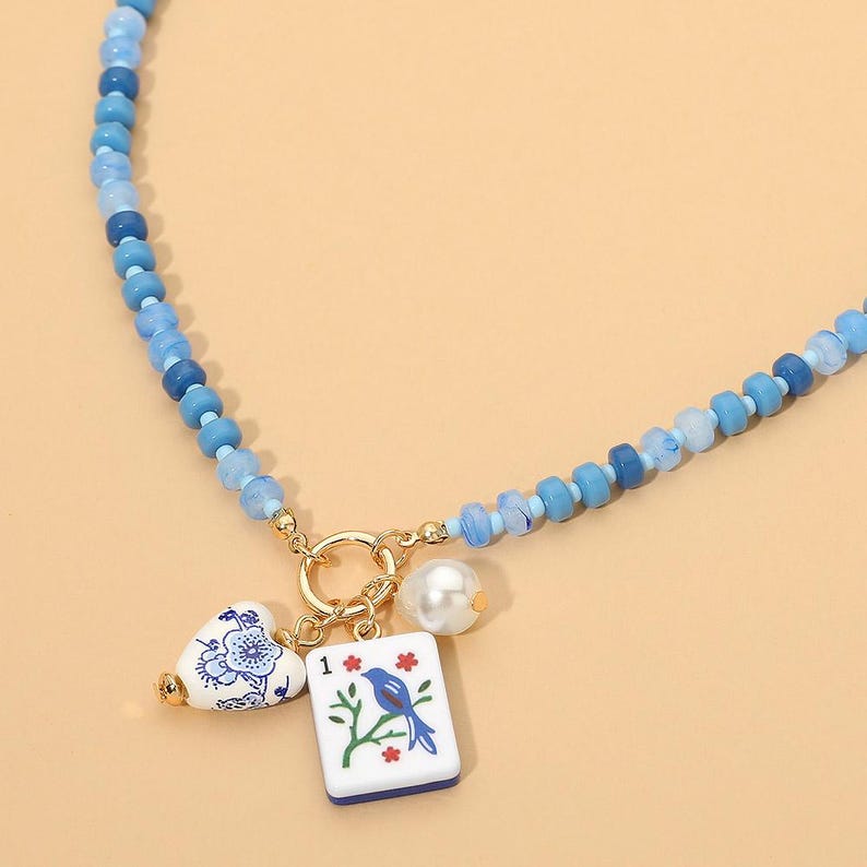 May include: A beaded necklace featuring various shades of blue beads and a gold-toned circular clasp. Three charms hang from the clasp: a white heart with blue floral patterns, a white rectangular mahjong tile with a blue bird, and a white pearl.
