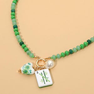 May include: A necklace featuring a strand of green beads in varying shades. The center includes a gold-toned ring holding three charms: a white heart with green floral patterns, a white pearl-like bead, and a white rectangular tile with a green bamboo design and the number 3.