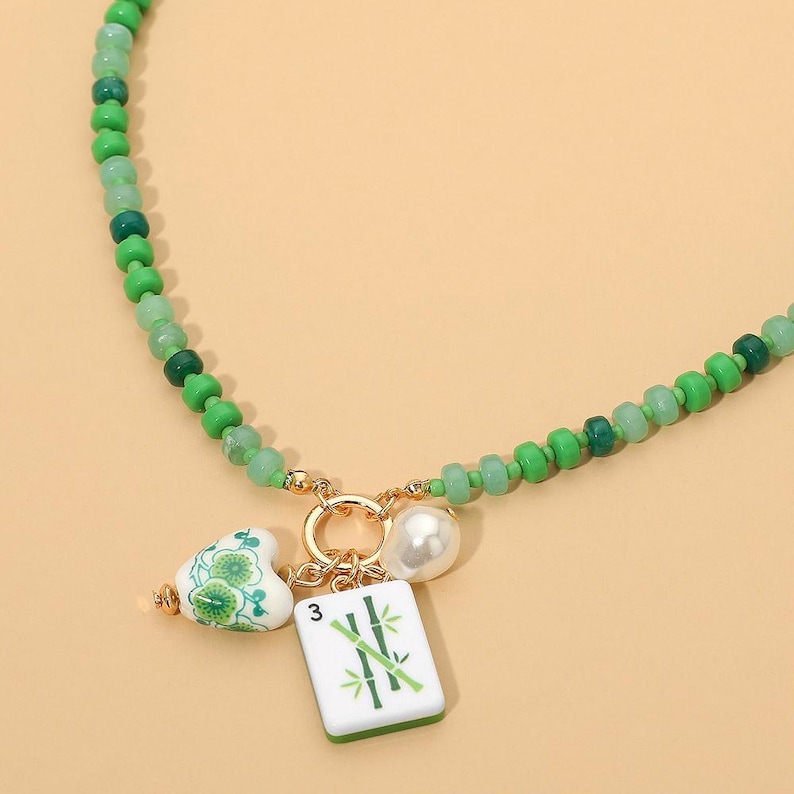 May include: A necklace featuring a strand of green beads in varying shades. The center includes a gold-toned ring holding three charms: a white heart with green floral patterns, a white pearl-like bead, and a white rectangular tile with a green bamboo design and the number 3.